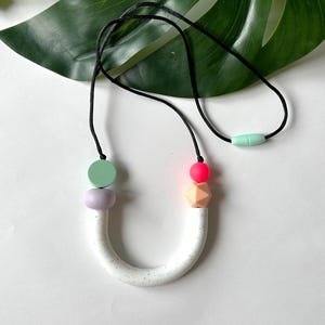 Peut inclure: Un collier en silicone blanc avec une finition mouchetée et un pendentif en forme de U. Le collier a un cordon noir et des perles en silicone colorées dans les tons vert, rose et orange.