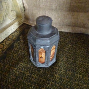 May include: A silver octagonal tea caddy with painted scenes on each side. The caddy is sitting on a patterned rug.