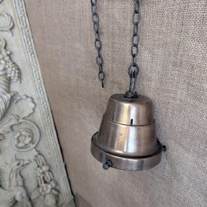 May include: A vintage-style brass pendant light fixture with a bell-shaped shade and a dark metal chain. The fixture is mounted on a textured, neutral-toned wall, showcasing its classic design and warm metallic finish.