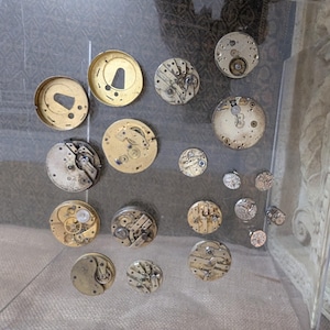 May include: A display of antique watch components, including cogs, plates, and casings, presented in a clear acrylic case. The parts vary in size and shape, with gold and silver hues, revealing intricate mechanical details.
