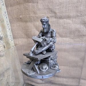May include: A detailed pewter sculpture of a seated man in historical attire, working with a hammer and chisel on a small anvil. The figure is on a small, round base with a hat and other details. The sculpture is approximately 6 inches tall.