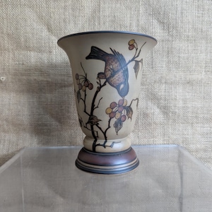 May include: A beige ceramic vase with a flared rim and a pedestal base. The vase features a detailed illustration of a bird perched on a branch with berries. The bird is brown and blue, and the berries are yellow and red.