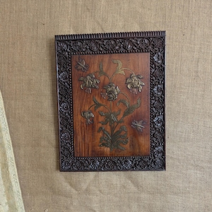 May include: A rectangular wooden wall art piece with a dark brown, ornate carved frame. The center features a floral design with brass accents on a brown wooden background. The artwork is mounted on a tan fabric.