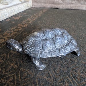 May include: A silver-colored turtle figurine with a detailed shell pattern. The figurine has its head and legs extended, giving it a lifelike appearance. The turtle is displayed on a patterned surface.