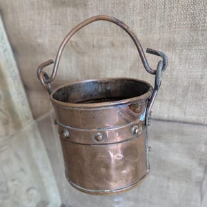 May include: A small, antique copper bucket with a curved handle. The bucket has a cylindrical body with a riveted band around the middle. The handle is attached with looped metal attachments. The copper has a warm, aged patina.