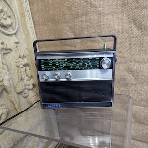 May include: A vintage Crown transistor radio with a black and silver fascia, tuning dials, and a carry handle. The radio has a black speaker grille and a blue "Crown" logo. It rests on a clear glass surface.