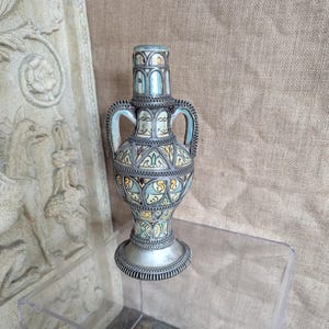 May include: An ornate, silver-toned vase with a detailed design. The vase features a bulbous body, two handles, and a tall neck. The design incorporates blue, yellow, and green accents. The vase sits on a clear display case.