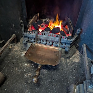 May include: A fireplace with burning coals and flames. A brass shovel with a wooden handle rests in front of the fireplace. The fireplace has a decorative metal grate.
