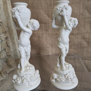 Könnte beinhalten: Zwei weiße Cherub-Statuen, die jeweils eine dekorative Vase halten. Die Cherubs stehen auf verzierten Sockeln mit wirbelnden Details. Die Vasen haben florale Akzente. Die Statuen werden auf einer transparenten Oberfläche vor neutralem Hintergrund ausgestellt.