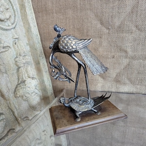May include: A silver-toned metal sculpture of a bird with a long neck and tail feathers, perched atop a turtle on a rectangular base. The bird holds a branch with small objects. The sculpture is displayed on a clear surface.