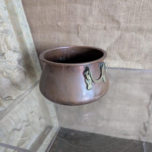 May include: A small, antique copper pot with brass handles. The pot has a rounded body and a wide opening. The handles are ornate and curved. The copper has a warm, reddish-brown colour, suggesting age and use. The pot is likely for decorative purposes.