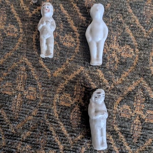 Puede incluir: Tres pequeñas muñecas antiguas de porcelana. Las muñecas son blancas con detalles pintados, incluyendo rasgos faciales. Las muñecas están de pie, con los brazos a los lados. Las muñecas están sobre una tela marrón y negra estampada.