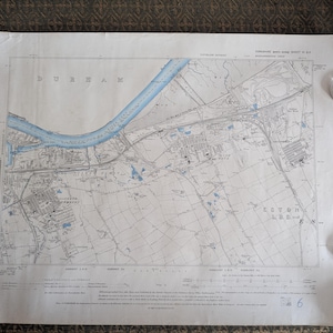 May include: A vintage map of Durham, England, with a blue river running through the center. The map is detailed with roads, buildings, and fields. Four silver coins are placed around the map's edges.