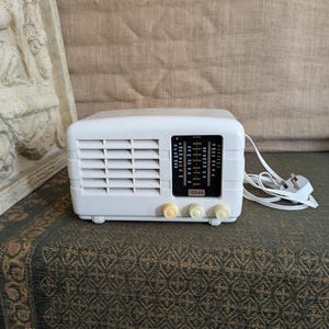 Może przedstawiać: Białe radio w stylu vintage o prostokątnym kształcie z kratką głośnika z przodu. Posiada czarną tarczę z oznaczeniami stacji i trzy kremowe pokrętła. Biały przewód zasilający z wtyczką wystaje z boku.