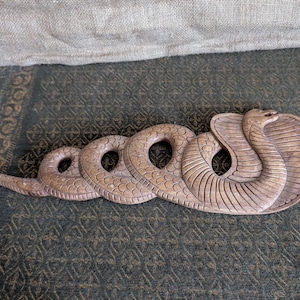 May include: A carved wooden cobra sculpture in shades of brown. The snake is depicted with its hood flared and body coiled. The sculpture has detailed scale and pattern carvings, and is lying on a patterned surface.