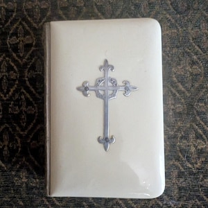 May include: Ivory-colored, rectangular book with a silver cross emblem. The book has a glossy finish and a silver border. The cross is detailed with ornate designs. The background is a dark, patterned fabric.