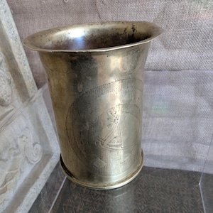 May include: An antique metal cup with a flared rim and a cylindrical body. The cup features an etched design of a figure within a circular pattern. The metal has a tarnished appearance, indicating age. The cup is likely an antique or a replica.