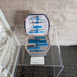 May include: Vintage manicure set with a silver-tone metal case and a light blue interior. The set contains scissors, nail files, and grooming tools, secured by blue straps. A small card is visible at the bottom.