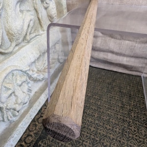 以下が含まれることがあります： 面取り加工が施された、木製のテーパーダウエル。ダウエルは明るい茶色で、濃い茶色の丸みを帯びた端があります。ダウエルは透明な表面に置かれています。