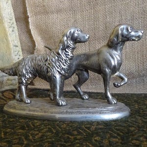 Puede incluir: Una figurita de plata de dos perros, uno de pie y otro acostado, sobre una base ovalada de plata.