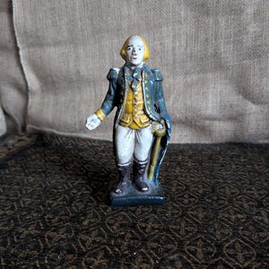 May include: A painted cast iron figurine of a man in a blue and yellow military uniform holding a staff. The figurine is standing on a black base.