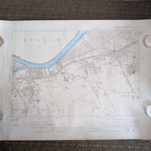 May include: A vintage map of Durham, England, with a blue river running through it. The map is held down by four silver coins. The word "DURHAM" is printed across the top of the map.