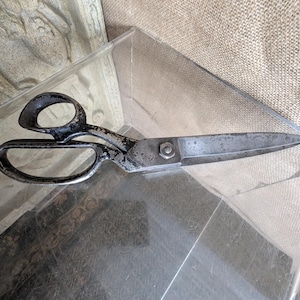 May include: A pair of vintage metal shears featuring a dark, weathered finish on the handles and blades. The tool includes two asymmetrical finger loops and a central metal nut securing the two blades together, showing signs of age and wear.