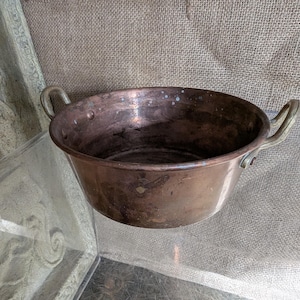 May include: A vintage copper bowl with two brass handles. The bowl has a wide, shallow shape and a rich, aged patina. The interior shows signs of use, adding to its antique charm. Ideal for kitchen decor.