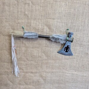 May include: A decorative tomahawk with a metal blade featuring a black silhouette design. The handle is wrapped with grey fur and adorned with white fringe. The tomahawk is a rustic, handcrafted item.