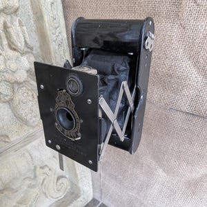 May include: A black folding camera with a bellows and a lens. The camera has a metal plate with the text "The No. 1 Kodak Folding Pocket Kodak" on it.