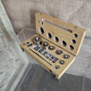 May include: A vintage wooden weight set with a hinged lid. The set includes various metal weights and small rectangular weights, arranged in a light-coloured wooden box. The box has several circular and rectangular openings.