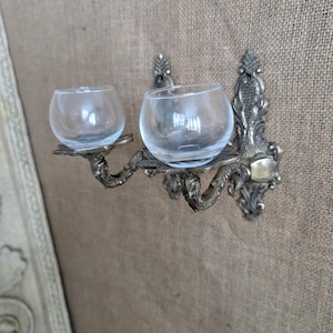 May include: A wall-mounted double candle holder featuring an ornate, antique-style brass base with intricate scrollwork and two curved arms. Each arm supports a clear, spherical glass cup designed to hold a small candle or tea light.