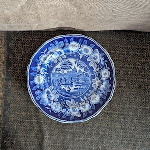 May include: A round ceramic plate featuring a blue and white transferware pattern. The center depicts a pastoral landscape with a river, a small boat, and a cottage. The wide border is decorated with detailed floral motifs and foliage.