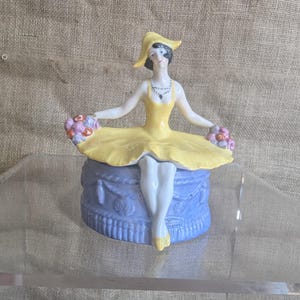 May include: A vintage porcelain figurine of a seated woman in a yellow dress and hat, holding bouquets. The figurine is perched atop a blue, cake-shaped base. The woman has black hair and a necklace. The figurine is approximately 15 cm tall.