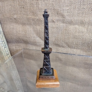 Puede incluir: Una réplica en miniatura de una torre alta y oscura con un diseño de celosía detallado. La torre está montada sobre una pequeña base de madera rectangular. La base tiene una pequeña placa de metal con texto. Es probable que sea un recuerdo o un objeto decorativo.