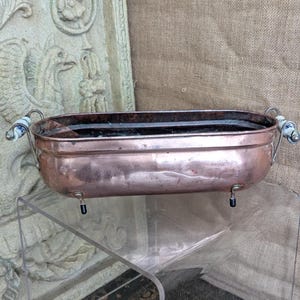 Könnte beinhalten: Ein länglicher Kupfer-Pflanzkübel mit polierter Metalloberfläche, der an beiden Seiten zwei geschwungene Metallgriffe mit weiß-blauen Keramikakzenten aufweist. Das Gefäß wird von vier kleinen, dunkel gefärbten Füßen an der Unterseite getragen.