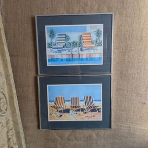 May include: Two framed prints of beach scenes. The top print shows two striped beach chairs by a swimming pool. The bottom print shows three striped beach chairs on a sandy beach.