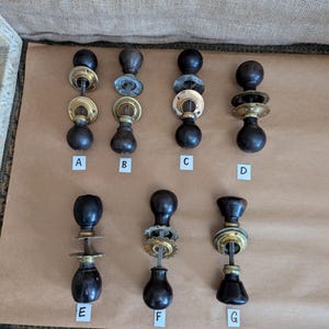 Op de afbeelding: Zeven antieke deurknoppen met donkere houten handvatten en messing of zilverkleurige metalen accenten zijn gerangschikt op een bruin oppervlak. Elke knop heeft een uniek ontwerp, dat vintage hardwarestijlen laat zien.