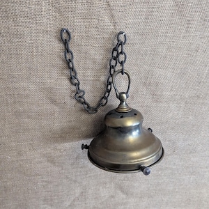 May include: An antique brass-colored pendant light fixture with a bell-shaped shade and a dark metal chain. The fixture has small decorative screws and a circular design. The chain is attached to the top of the fixture.