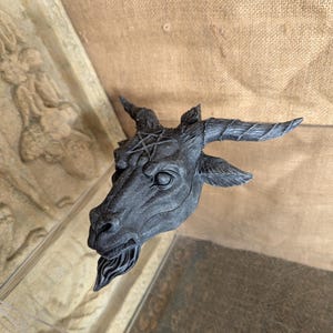 May include: A dark gray sculpture of a goat's head with curved horns and a pentagram on its forehead. The goat has a detailed beard and is set against a neutral background. The sculpture is likely a decorative item.