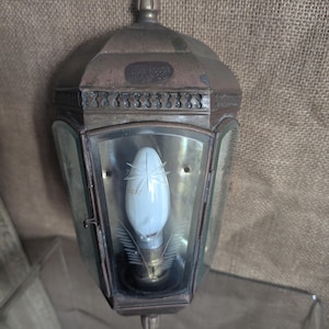 May include: A wall-mounted lantern with a weathered bronze-toned metal frame and clear glass panels. The metal top features the embossed text LIME HOUSE LAMP CO. A single white light bulb is visible inside, featuring etched star and leaf designs.