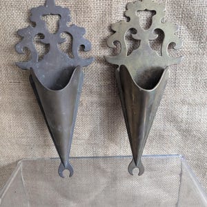 May include: Two antique metal wall sconces. Each sconce has a decorative top with a stylized floral design and a cone-shaped holder. The sconces are made of a dark metal with a slightly aged appearance, and they have a small hook at the bottom.