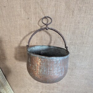 May include: A rustic copper cauldron with a curved handle. The cauldron has a rounded body and a slightly flared rim. The metal has a weathered appearance with areas of green oxidation, suggesting age and use. The handle is made of iron.