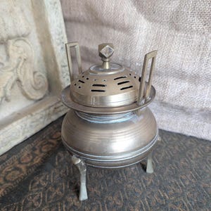 May include: An antique brass incense burner with a domed lid and ventilation holes. The burner has a round base supported by three small legs and two handles. The design is ornate and detailed, with a vintage aesthetic. The burner is made of brass.