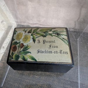 May include: A rectangular black box with a floral design and the text "A Present From Stockton-on-Tees". The box has a decorative border and is set inside a clear plastic container.