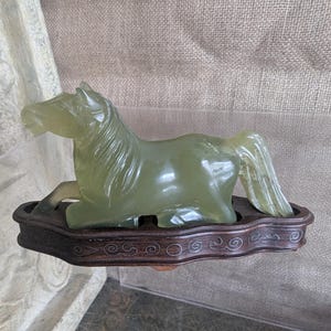 Puede incluir: Una escultura de caballo de jade translúcido en posición reclinada, montada sobre una base de madera oscura con tallas decorativas. El caballo es de color verde pálido y la base es de color marrón oscuro. Es un objeto decorativo.
