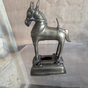 Puede incluir: Pequeña figura de caballo de latón antiguo. El caballo está de pie sobre una base rectangular con una silla de montar y una brida. La figura tiene un diseño detallado y es probablemente un artículo decorativo.