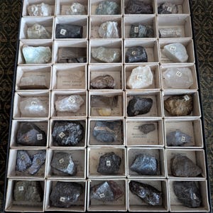 May include: A collection of rocks and minerals in a vintage box, each in its own compartment. The specimens are labelled with numbers. The rocks vary in colour and texture, including white, grey, and black stones.
