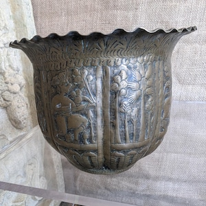 Puede incluir: Un macetero grande, ornamentado, de bronce con un diseño en relieve de palmeras y figuras. El macetero tiene un borde festoneado y una base redondeada.