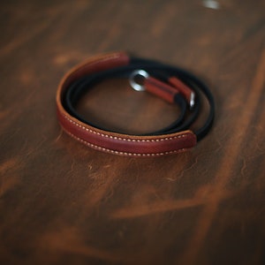 May include: A brown leather camera strap with a black leather lining and a silver metal loop.
